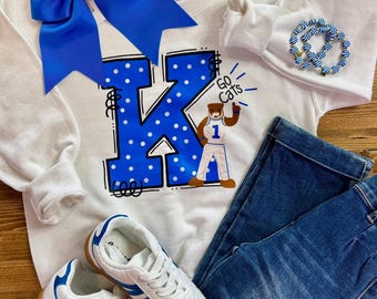 May include: A white sweatshirt with a large blue letter "K" with white polka dots and the words "Go Cats". The sweatshirt is paired with blue jeans, white sneakers with blue stripes, a blue bow, and a blue and white beaded bracelet.