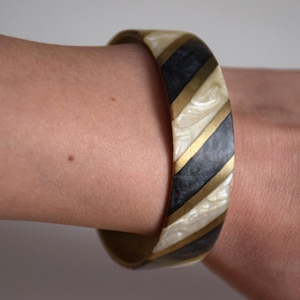 Könnte beinhalten: Ein breites Armreif-Armband mit einem schwarz-weißen Streifenmuster. Die Streifen sind mit goldenen Verzierungen versehen.