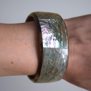 Op de afbeelding: Een houten armband met een grote, iriserende inleg van parelmoer. De inleg is gemaakt van kleine, onregelmatige stukjes schelp die een mozaïekpatroon creëren.