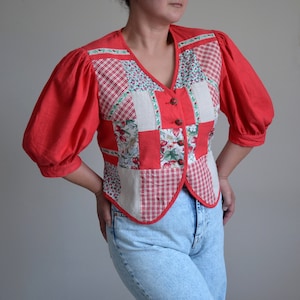 Puede incluir: Una blusa de color rojo, blanco y verde con estampado de patchwork y mangas abullonadas. La blusa tiene cuello en V y cierre de botones.