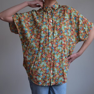 May include: A patterned button-down shirt with a floral design. The shirt has a collar and short sleeves. The floral pattern includes red, blue, and yellow flowers on a brown base. The shirt is worn with blue jeans.