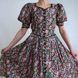 Könnte beinhalten: Ein schwarzes Kleid mit einem lebendigen Blumenmuster in Rosa, Rot, Gelb und Weiß. Das Kleid hat kurze Puffärmel, einen eckigen Ausschnitt, ein tailliertes Oberteil mit einer schwarzen Knopfleiste auf der Vorderseite und einen Faltenrock.