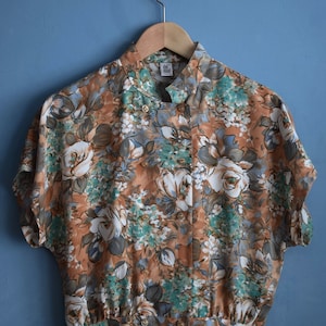 May include: A brown, short-sleeved blouse with a floral print featuring white, green, and blue flowers. The blouse has a mandarin collar and a button-up front. The label "Del Mod" is visible on the front of the blouse.