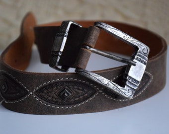 May include: A brown leather belt with a silver buckle featuring an intricate design. The belt has a stitched pattern of diamond shapes.