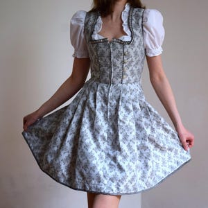 May include: A traditional German dirndl dress. The dress features a light blue floral patterned skirt and a gray bodice with decorative buttons. The white blouse has short, puffed sleeves. The dress has a gray trim.