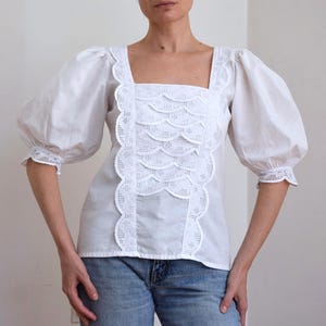 May include: A white blouse featuring voluminous puff sleeves with lace-trimmed cuffs, a square neckline, and a central panel decorated with scalloped eyelet embroidery. The garment is paired with blue denim jeans, creating a clean, neutral aesthetic.