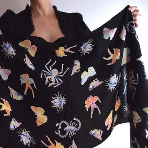 Op de afbeelding: Zwarte sjaal met een kleurrijk insecten- en vlinderpatroon. De sjaal heeft verschillende insecten en vlinders in tinten oranje, geel, blauw en roze. De sjaal is gemaakt van een zacht materiaal.