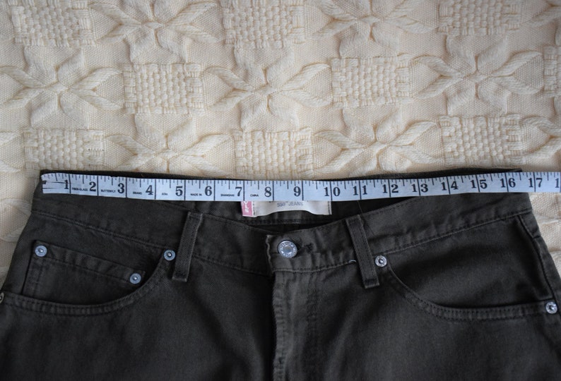 May include: A pair of dark green jeans with a measuring tape across the waistband. The tape reads 17 inches.