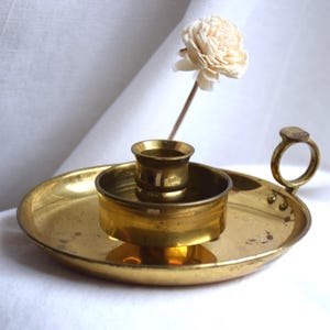 Puede incluir: Un candelero de latón vintage con base circular y asa pequeña. El candelero tiene una copa central para la vela y un plato circundante. Una flor decorativa se coloca detrás del candelero.
