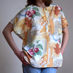 May include: A short-sleeved blouse with a colorful pattern featuring red cherries, brown leaves, and abstract shapes in shades of yellow, orange, and blue. The blouse has a round neckline and a single button closure.