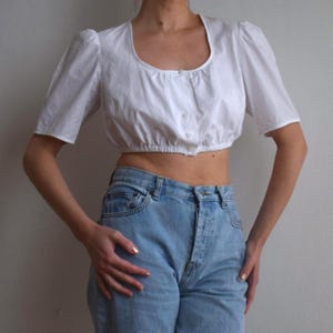 Puede incluir: Una blusa blanca corta con mangas cortas abullonadas y un escote cuadrado. La blusa tiene una botonadura frontal y está fruncida en la cintura. El modelo lleva vaqueros azul claro.