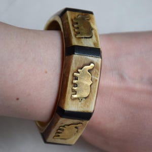 May include: A square, ivory-colored bracelet with gold elephant designs and black accents. The bracelet is worn on a wrist and has a geometric shape. The elephant designs are detailed and add a unique touch to the accessory.