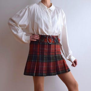 May include: A vintage outfit comprising a white blouse with a lace collar and a long-sleeved design, paired with a red, black, and green plaid pleated skirt. The skirt is cinched with a wide belt and a silver-tone buckle. A classic, preppy ensemble.