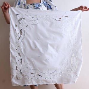 Pode incluir: Uma toalha de mesa quadrada branca com bordados florais intrincados e detalhes vazados nas bordas. O tecido é segurado, mostrando o design delicado e a textura. É um item vintage ou antigo.
