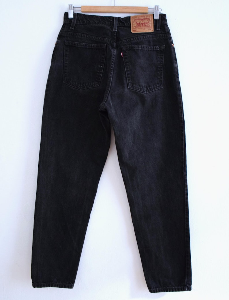 Peut inclure: Un jean en denim noir, suspendu &agrave; un cintre en bois. Le jean a deux poches arri&egrave;re et une &eacute;tiquette rouge et blanche. Le denim semble l&eacute;g&egrave;rement d&eacute;lav&eacute;.