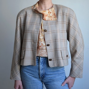Puede incluir: Una chaqueta de cuadros gris y marrón recortada con cierre de botones y bolsillos de solapa. La chaqueta tiene cuello y se lleva sobre una blusa con estampado floral.