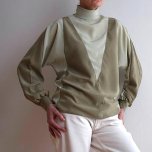 Può includere: Una camicetta a maniche lunghe nei toni del verde oliva e verde chiaro. La camicetta presenta un collo alto a lupetto e un design a forma di V sul davanti. Le maniche sono lunghe e raccolte ai polsi. La persona indossa pantaloni color crema.