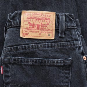 Peut inclure: Gros plan sur un jean en denim noir d'aspect d&eacute;lav&eacute;. La poche arri&egrave;re comporte une petite &eacute;tiquette rouge. Un patch en cuir beige avec des lettres rouges et un logo de chariot tir&eacute; par des chevaux est visible. Le jean est &eacute;tiquet&eacute; "550".