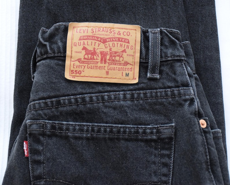 Peut inclure: Gros plan sur un jean en denim noir d'aspect d&eacute;lav&eacute;. La poche arri&egrave;re comporte une petite &eacute;tiquette rouge. Un patch en cuir beige avec des lettres rouges et un logo de chariot tir&eacute; par des chevaux est visible. Le jean est &eacute;tiquet&eacute; "550".