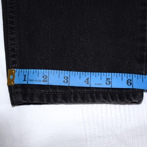 Peut inclure: Gros plan sur un jean en denim noir avec un m&egrave;tre ruban bleu indiquant une largeur d'environ 17,8 cm. Le jean est pli&eacute; &agrave; l'ourlet, et le m&egrave;tre ruban est plac&eacute; au bas du jean.