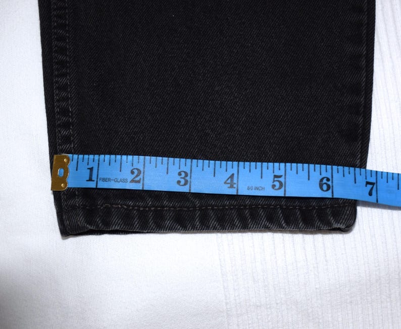Peut inclure: Gros plan sur un jean en denim noir avec un m&egrave;tre ruban bleu indiquant une largeur d'environ 17,8 cm. Le jean est pli&eacute; &agrave; l'ourlet, et le m&egrave;tre ruban est plac&eacute; au bas du jean.