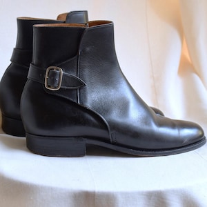 Puede incluir: Un par de botines de cuero negro. Los botines tienen un tacón bajo, una correa con hebilla plateada y un acabado liso y pulido. Los botines se exhiben sobre una superficie blanca.