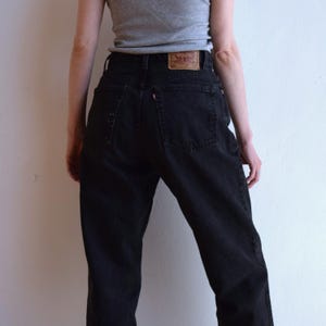 Peut inclure: Jean noir taille haute avec un design classique &agrave; cinq poches. Le jean pr&eacute;sente un patch en cuir au dos avec un logo de marque. La personne porte un d&eacute;bardeur gris.