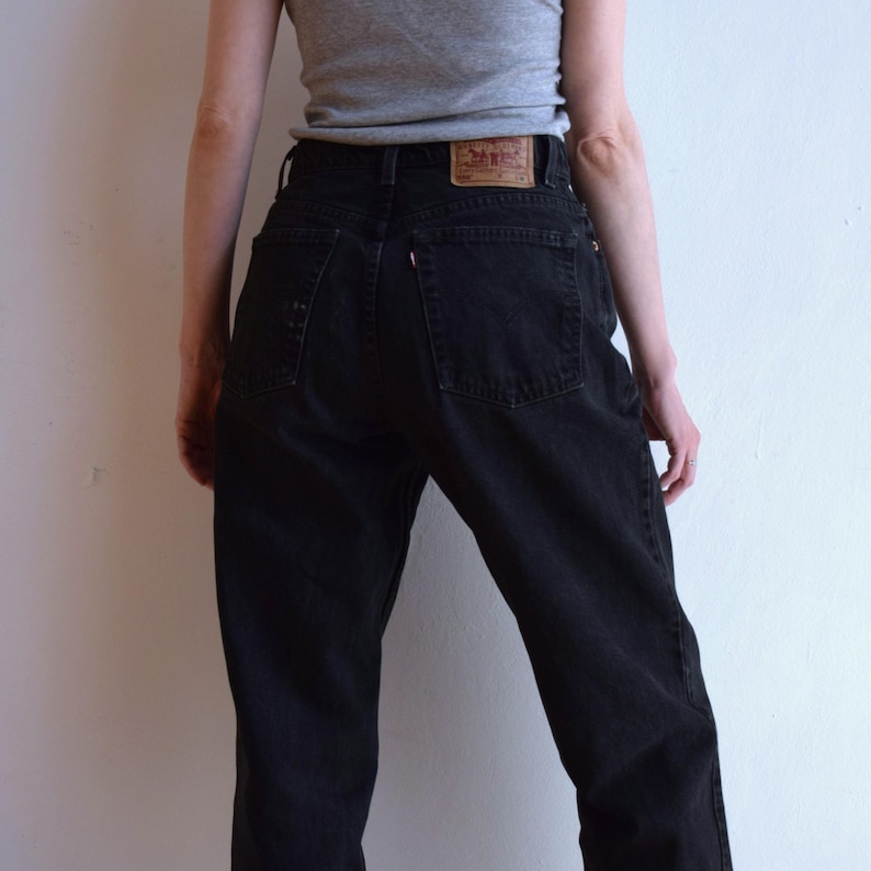 Peut inclure: Jean noir taille haute avec un design classique &agrave; cinq poches. Le jean pr&eacute;sente un patch en cuir au dos avec un logo de marque. La personne porte un d&eacute;bardeur gris.