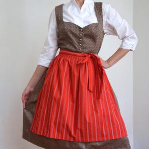 May include: A white blouse with three-quarter sleeves worn under a brown bodice featuring a small repeating diamond pattern and metal buttons. A bright red apron with vertical white stripes and a tied waist sits over a matching brown skirt.