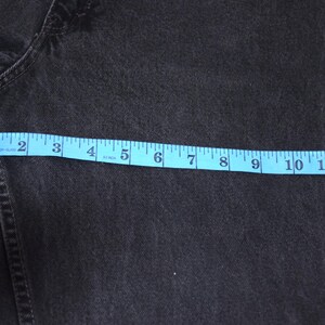Peut inclure: Gros plan sur un jean en denim noir avec un ruban &agrave; mesurer bleu indiquant une mesure d'environ 33 cm. Le jean est de couleur fonc&eacute;e et semble &ecirc;tre fait d'un mat&eacute;riau robuste.