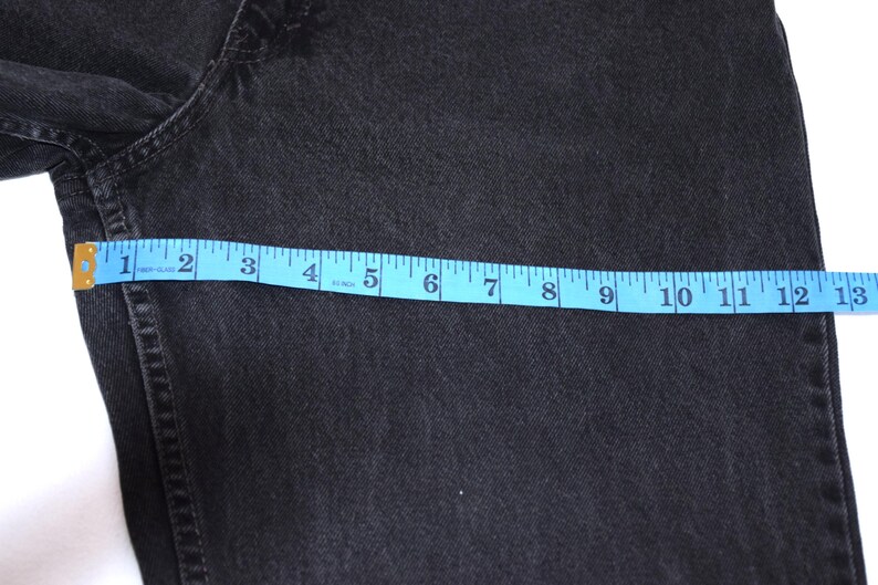 Peut inclure: Gros plan sur un jean en denim noir avec un ruban &agrave; mesurer bleu indiquant une mesure d'environ 33 cm. Le jean est de couleur fonc&eacute;e et semble &ecirc;tre fait d'un mat&eacute;riau robuste.