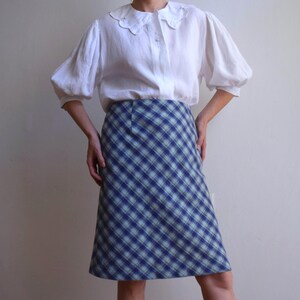 Könnte beinhalten: Ein Outfit im Vintage-Stil mit einer weißen Bluse mit Rüschenkragen und Puffärmeln, kombiniert mit einem blau-grünen karierten Rock. Der Rock reicht bis zu den Knien und erzeugt eine klassische Retro-Ästhetik.