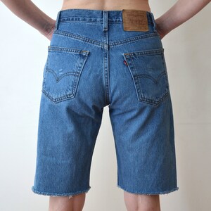 Puede incluir: Un par de pantalones cortos de mezclilla azul con dobladillo deshilachado. Los pantalones cortos tienen un diseño clásico de cinco bolsillos y un parche de cuero marrón en la parte posterior de la cintura.