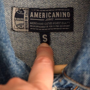Może przedstawiać: Niebieska kurtka dżinsowa z metką, na kt&oacute;rej widnieje napis "AMERICANINO JEANS" i "STYLED IN ITALY". Metka zawiera r&oacute;wnież napis "AMERICANINO CLOTHES HIGHEST QUALITY PROPORTIONED FIT FOR EXTRA COMFORT". Metka rozmiaru wskazuje rozmiar "S".