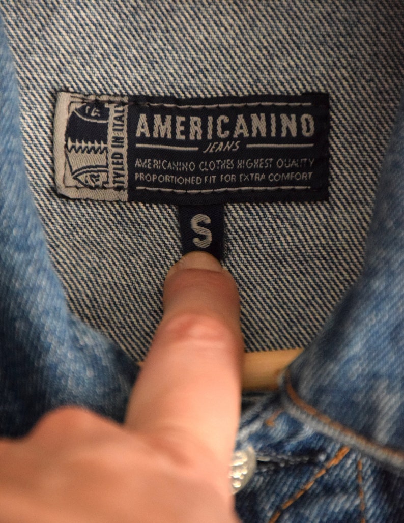 Może przedstawiać: Niebieska kurtka dżinsowa z metką, na kt&oacute;rej widnieje napis "AMERICANINO JEANS" i "STYLED IN ITALY". Metka zawiera r&oacute;wnież napis "AMERICANINO CLOTHES HIGHEST QUALITY PROPORTIONED FIT FOR EXTRA COMFORT". Metka rozmiaru wskazuje rozmiar "S".