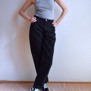 Peut inclure: Une personne porte un d&eacute;bardeur gris, un jean noir et des baskets noires et blanches. Le jean est taille haute et les baskets ont des lacets blancs. La personne se tient debout, les bras le long du corps.