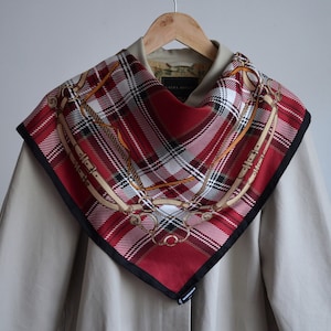 May include: A red and white plaid scarf with a black border and gold chain design. The scarf is draped over a beige jacket.