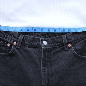 Peut inclure: Gros plan sur un jean en denim noir avec un ruban &agrave; mesurer bleu sur la taille. Le jean a des rivets en cuivre et une fermeture boutonn&eacute;e. La taille mesure environ 33 cm.