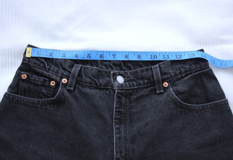 Peut inclure: Gros plan sur un jean en denim noir avec un ruban &agrave; mesurer bleu sur la taille. Le jean a des rivets en cuivre et une fermeture boutonn&eacute;e. La taille mesure environ 33 cm.