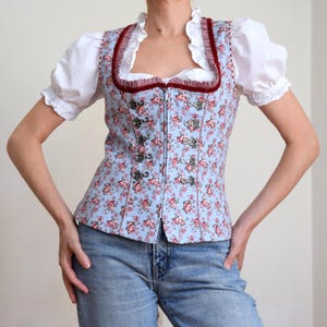 Pode incluir: Um corpete azul claro com um pequeno padrão floral vermelho e branco, decote quadrado com folhos brancos e uma borda de veludo vermelho escuro. O fecho frontal inclui ganchos de metal prateado e um zíper, usado sobre uma blusa branca.