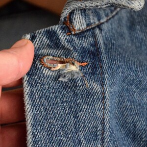 Puede incluir: Primer plano de una chaqueta vaquera azul con un peque&ntilde;o agujero e hilos deshilachados cerca del cuello.