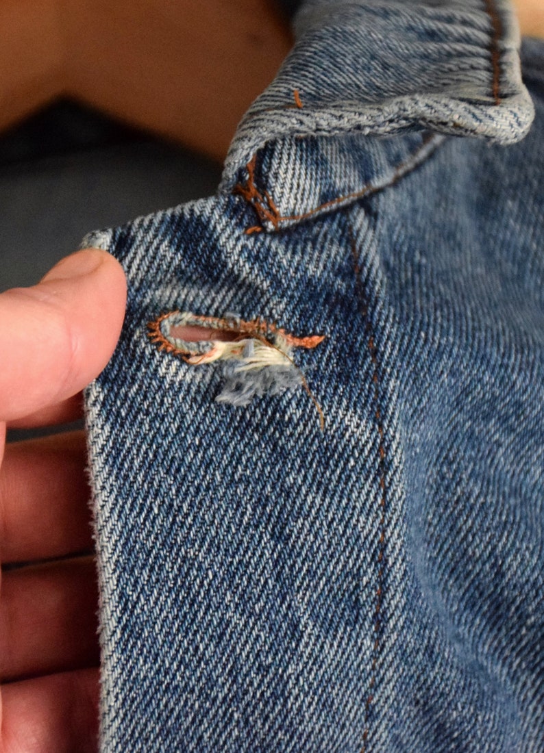 Puede incluir: Primer plano de una chaqueta vaquera azul con un peque&ntilde;o agujero e hilos deshilachados cerca del cuello.