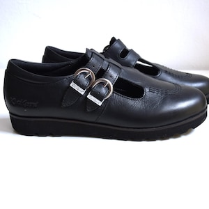 May include: Black leather buckle shoes with a strap across the top and a cut-out detail. The shoes have a thick sole.