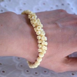 Puede incluir: Un brazalete blanco de cuentas redondas pequeñas. Las cuentas están dispuestas en un patrón que se asemeja a una flor.