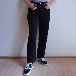 Peut inclure: Un jean noir taille haute avec une coupe droite. Le jean est associ&eacute; &agrave; des baskets noires et blanches. La personne porte un t-shirt gris. L'arri&egrave;re-plan est un mur blanc uni et un sol en bois.