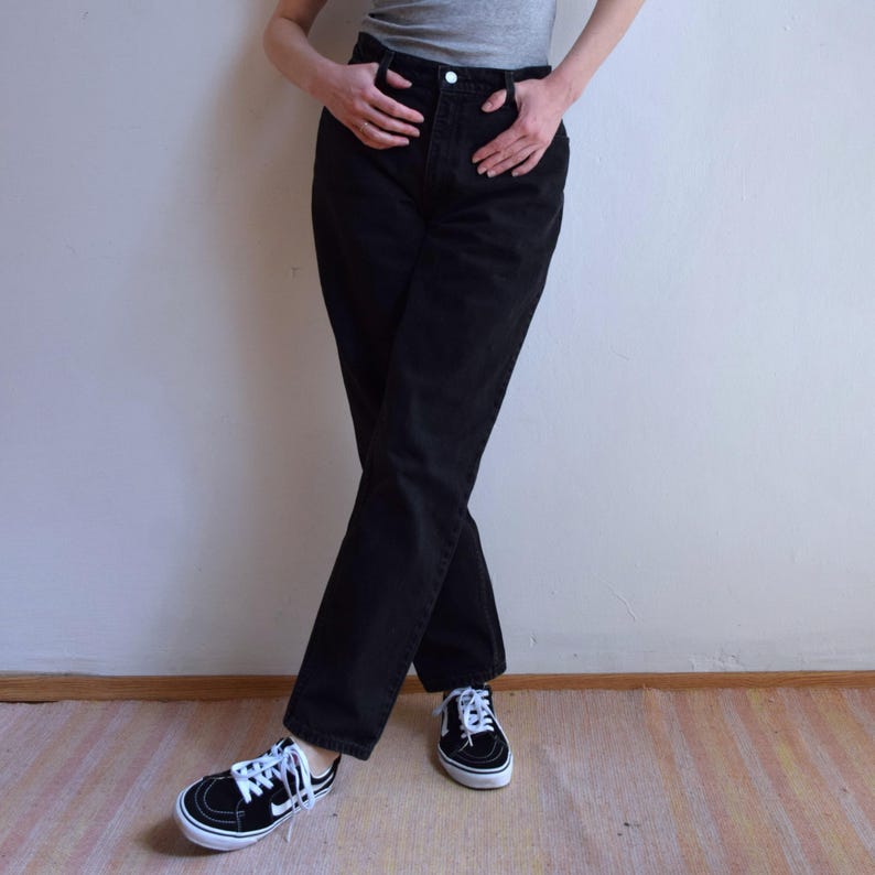 Peut inclure: Un jean en denim noir taille haute et coupe droite. Le jean est associ&eacute; &agrave; des baskets noires et blanches. La personne porte un d&eacute;bardeur gris. L'arri&egrave;re-plan est un mur blanc uni et un sol en bois.
