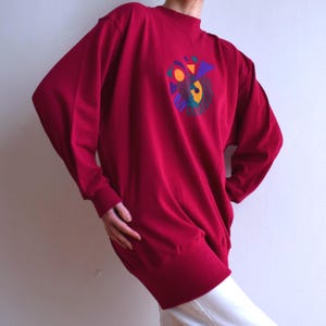 May include: A burgundy long-sleeved sweatshirt with a high neckline. The sweatshirt features a colourful geometric design on the chest, including orange, purple, and green shapes. The sleeves are long and the cuffs are ribbed.