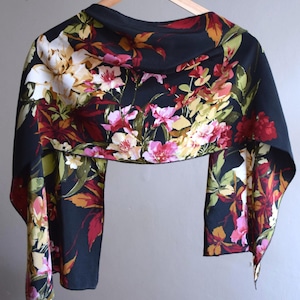 May include: A black shawl with a vibrant floral pattern. The design features a variety of flowers in shades of pink, white, yellow, and red, set against a dark background. The shawl is draped over a wooden hanger.