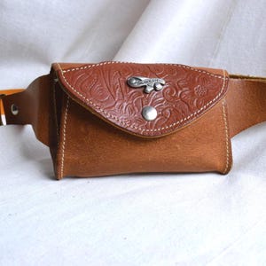 May include: A brown leather fanny pack with a floral embossed flap and a silver-tone clasp. The pack features a stitched design and an adjustable strap. The flap is secured with a silver snap. The text "OIL HOLSTER" is visible on the clasp.