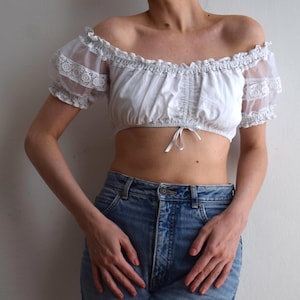 Può includere: Un top corto bianco con spalle scoperte, caratterizzato da maniche a sbuffo trasparenti con dettagli in pizzo. Il capo presenta uno scollo arricciato, un pannello frontale pieghettato e una piccola chiusura a laccio sull'orlo, abbinato a jeans blu.